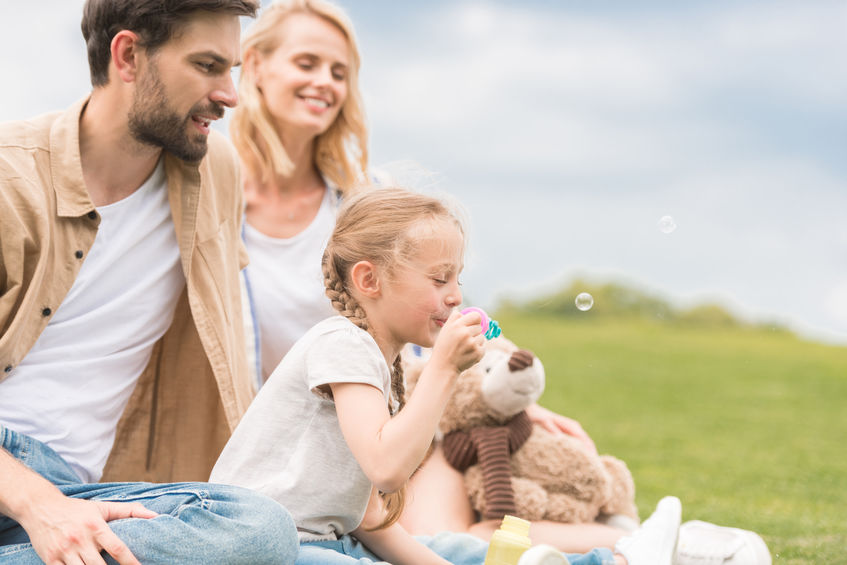 happy parents looking at cute little daughter with teddy bear blowing soap bubbles in park